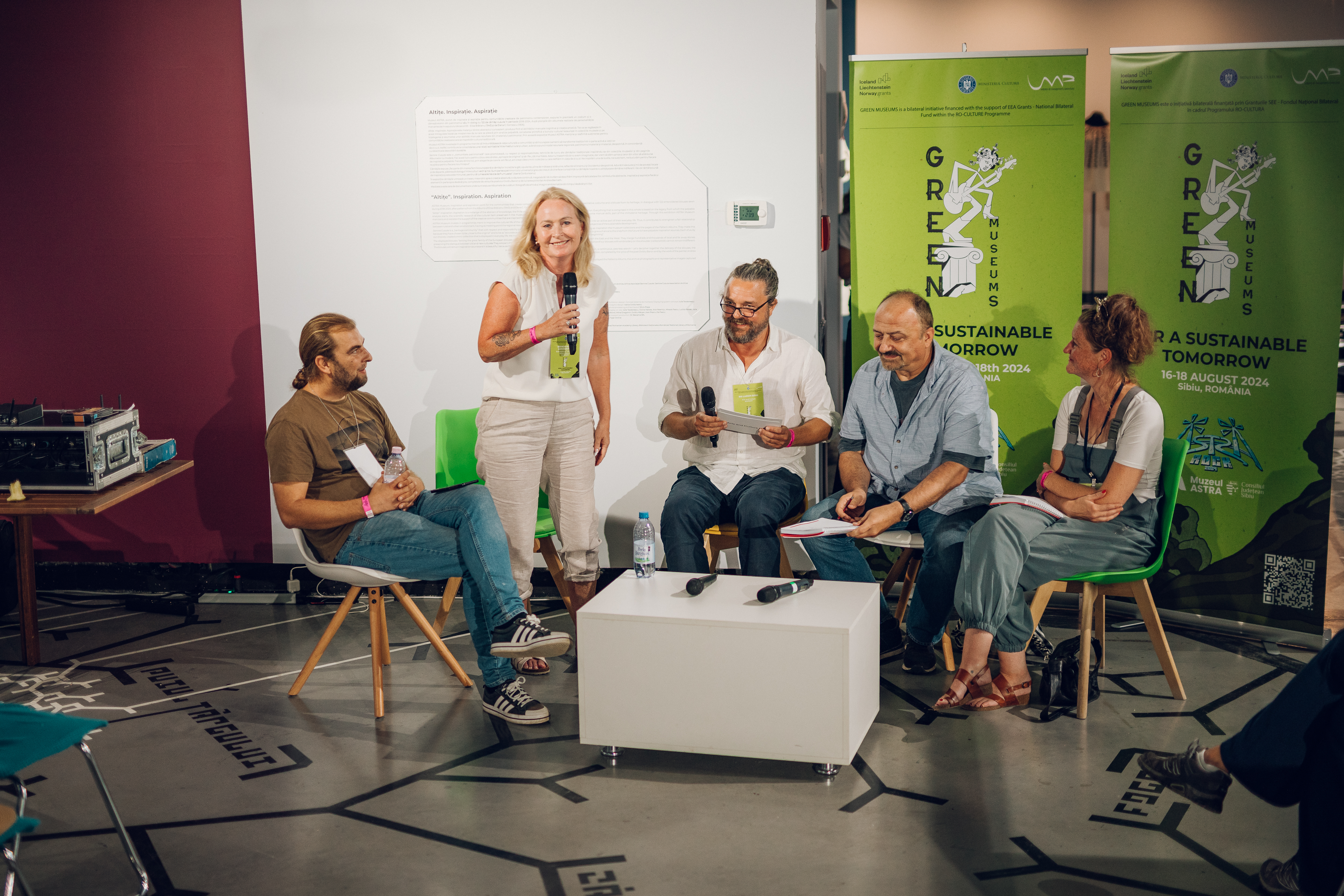 Fem voksne deltar i en paneldiskusjon eller presentasjon innendørs. De sitter rundt et lite hvitt bord med mikrofoner og vannflasker. Én kvinne står og snakker i mikrofon, mens de andre lytter eller forbereder seg på å snakke. Bak dem står to grønne bannere med teksten 'GREENHOUSE' og 'A SUSTAINABLE TOMORROW', samt datoen '18–20 AUGUST 2022'. Rommet har geometrisk gulvdesign, hvit vegg med termostat og en burgunderrød veggseksjon. Bildet viser et øyeblikk fra en konferanse med fokus på bærekraft.