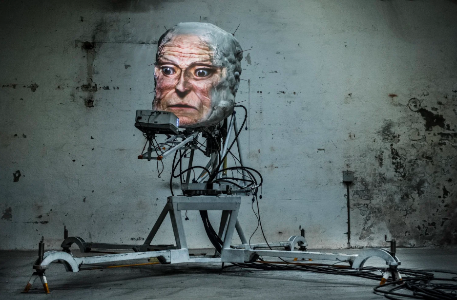 Kunstverket Requiem for an Exit (2024) av Thomas Kvam og Frode Oldereid: et stort, realistisk menneskehode montert på en mekanisk konstruksjon av metall, kabler og elektronikk, plassert i et industrielt rom.