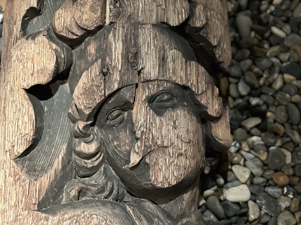 Nærbilde av en utskåret treskulptur som forestiller et menneskeansikt med stilisert hår og hjelm eller hodeplagg. Treverket har synlig struktur og sprekker, noe som tyder på alder eller slitasje. Bakgrunnen består av små, fargerike steiner, og antyder at skulpturen står utendørs, kanskje som del av en utstilling eller arkeologisk område. Bildet fremhever håndverkets detaljer og den historiske eller kunstneriske verdien.