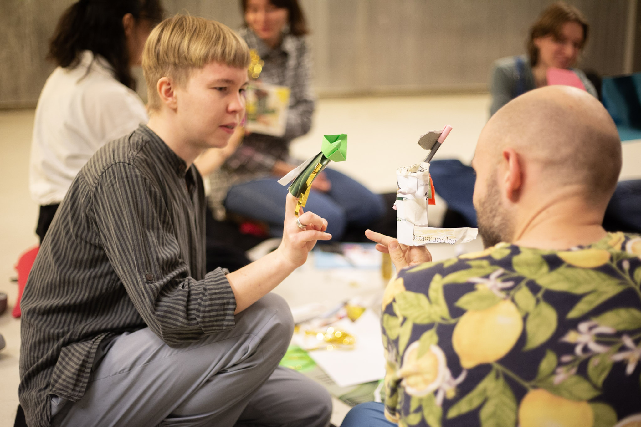 To personer sitter på gulvet og holder opp hver sin enkle figur laget av papir og papp, som de viser til hverandre. I bakgrunnen sitter flere deltakere samlet i en workshop-situasjon.