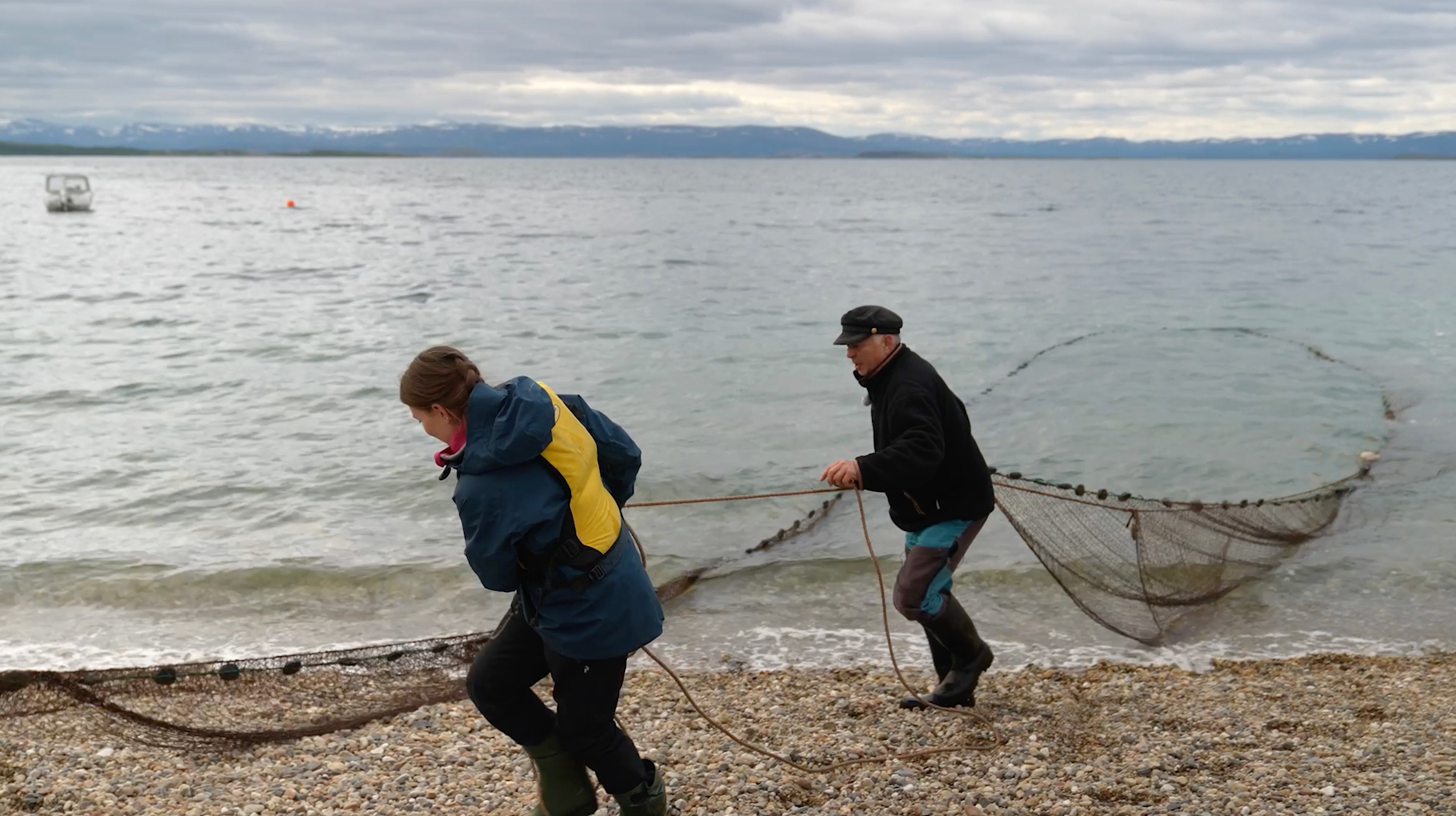 To personer står på en steinete strand og trekker et stort fiskegarn opp fra sjøen. Havet er rolig og himmelen er overskyet.