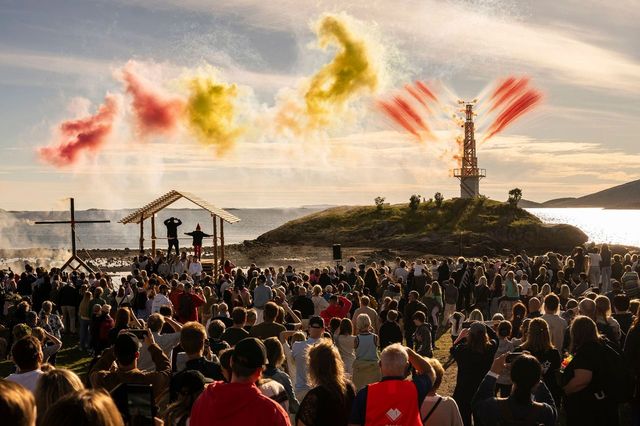 Stor folkemengde samlet ved en kystscene i kveldssol. Foran publikum står aktører under et åpent tretak, mens røyk i gule og røde farger skytes opp fra en installasjon på en liten holme ute i vannet. Hav, øyer og lav kveldssol danner bakgrunnen.