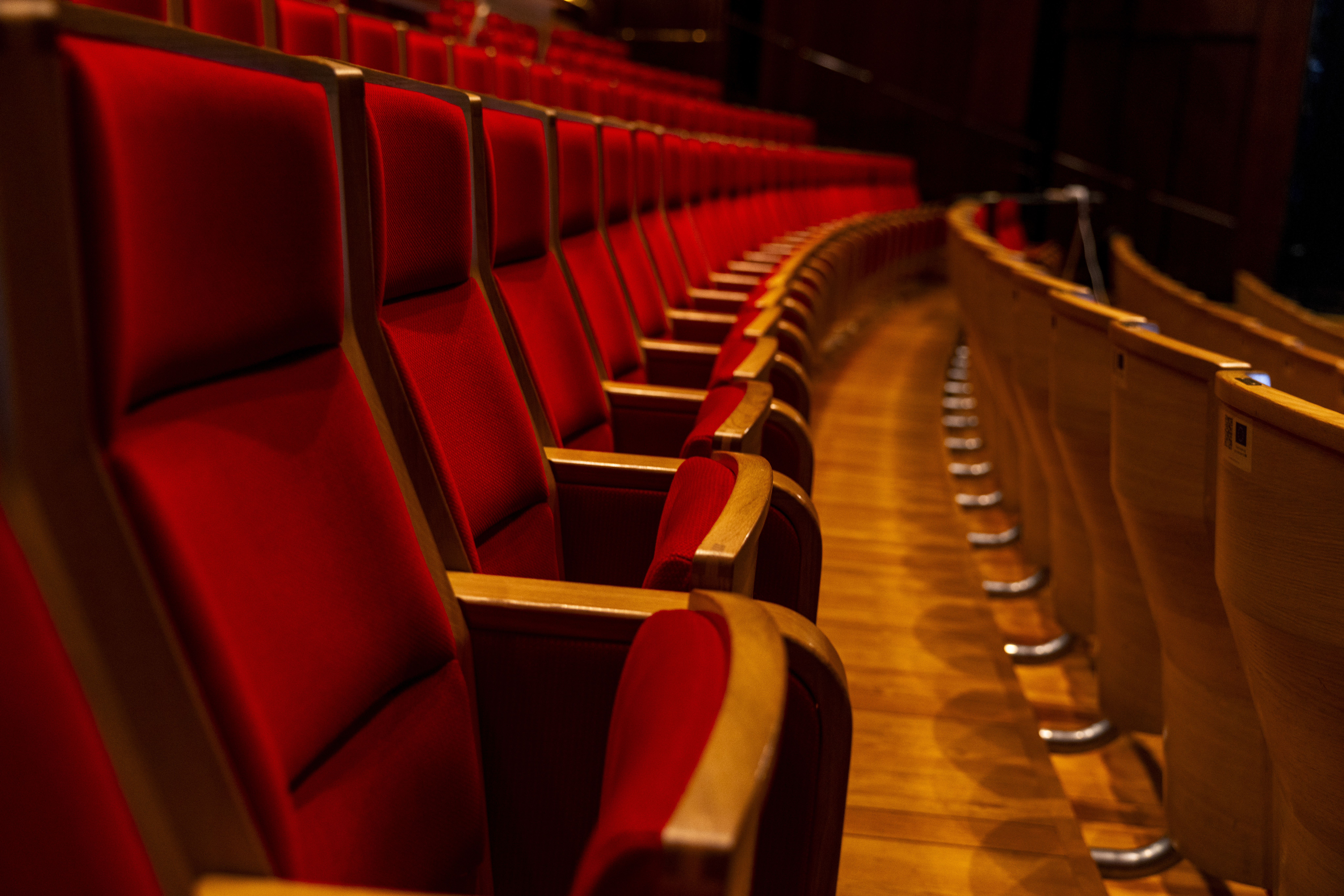 Rad med røde, polstrede teaterseter i en sal med buet oppsett og dempet belysning.