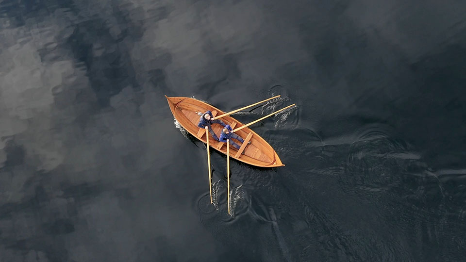 To personer ror en trebåt på stille, mørkt vann, sett ovenfra.