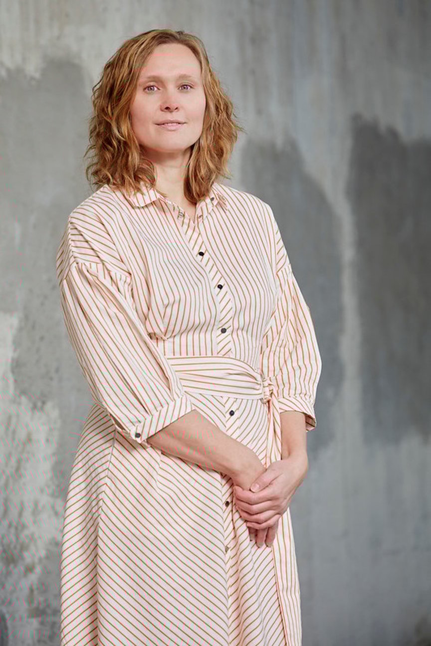 En kvinne står foran en grå vegg iført en hvit- og oransjestripete kjole. Hun har halvlangt lyst hår og hendene foldet foran kroppen.