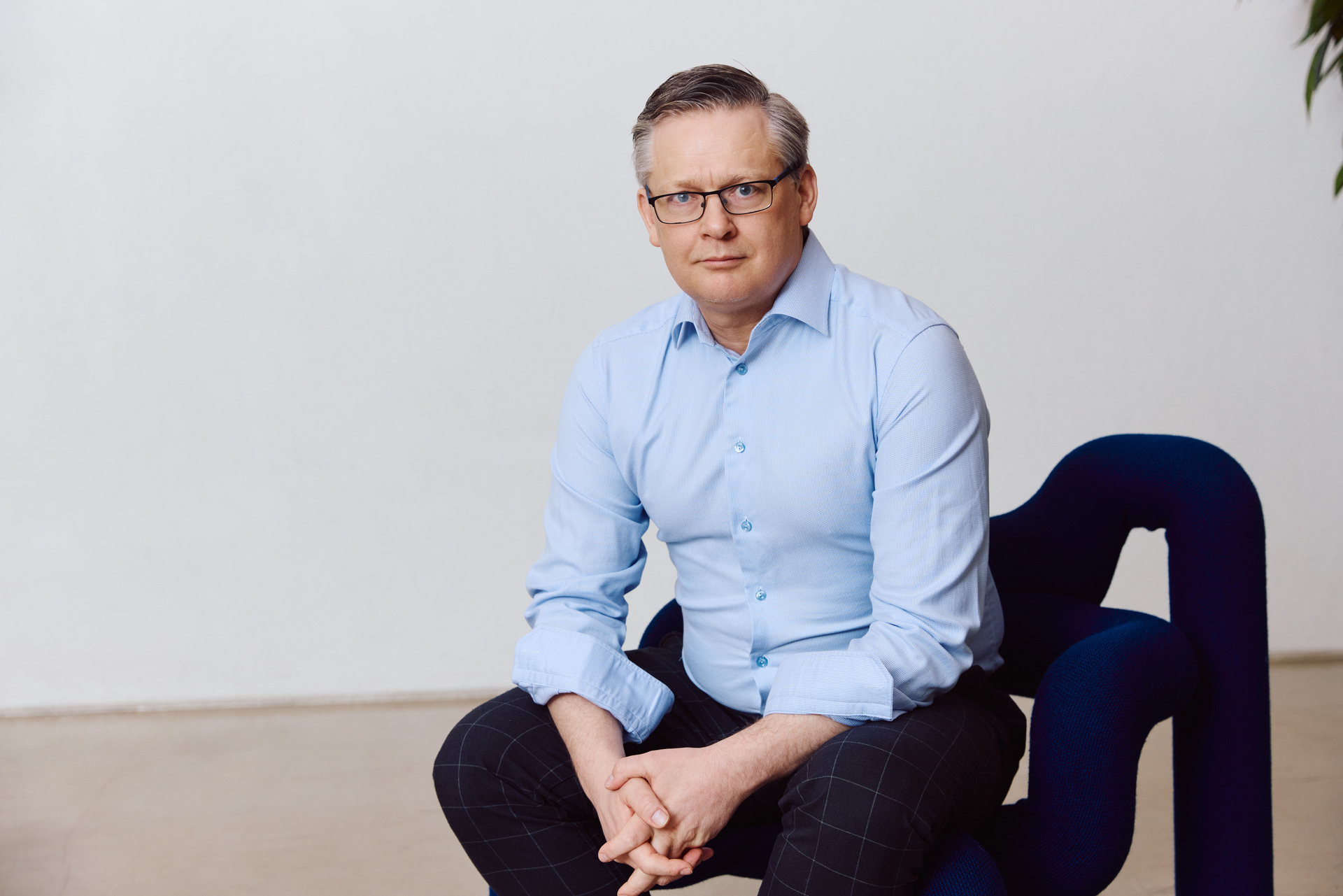 En middelaldrende mann med briller sitter på en blå stol, iført en lyseblå skjorte og mørke rutete bukser.