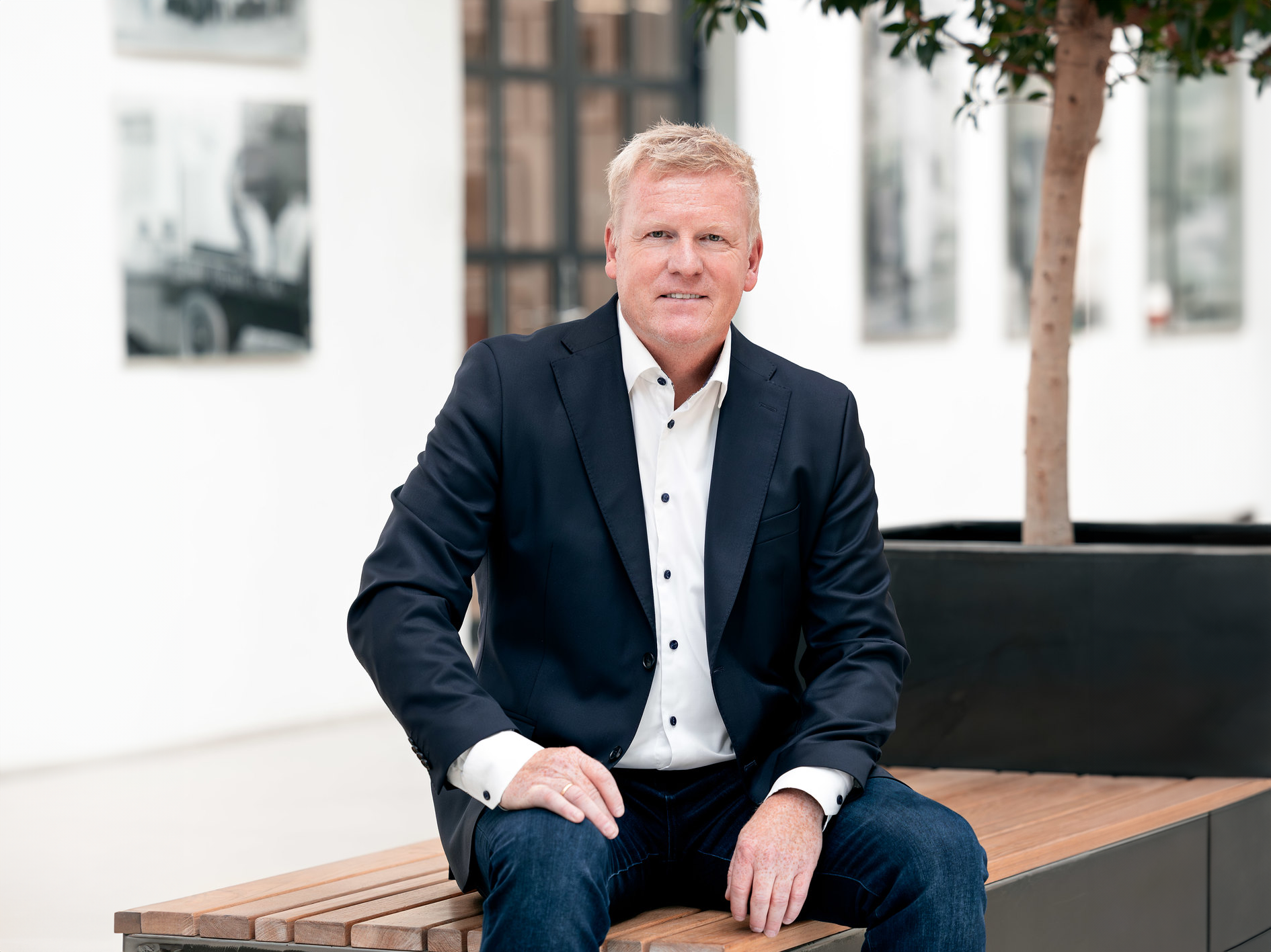 Øystein Strand ikledd en svart blazer, hvit skjorte og olabukser, sitter på en benk foran en plante.