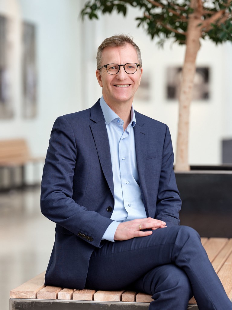 En mann iført mørkeblå dress sitter og smiler mot kamera.