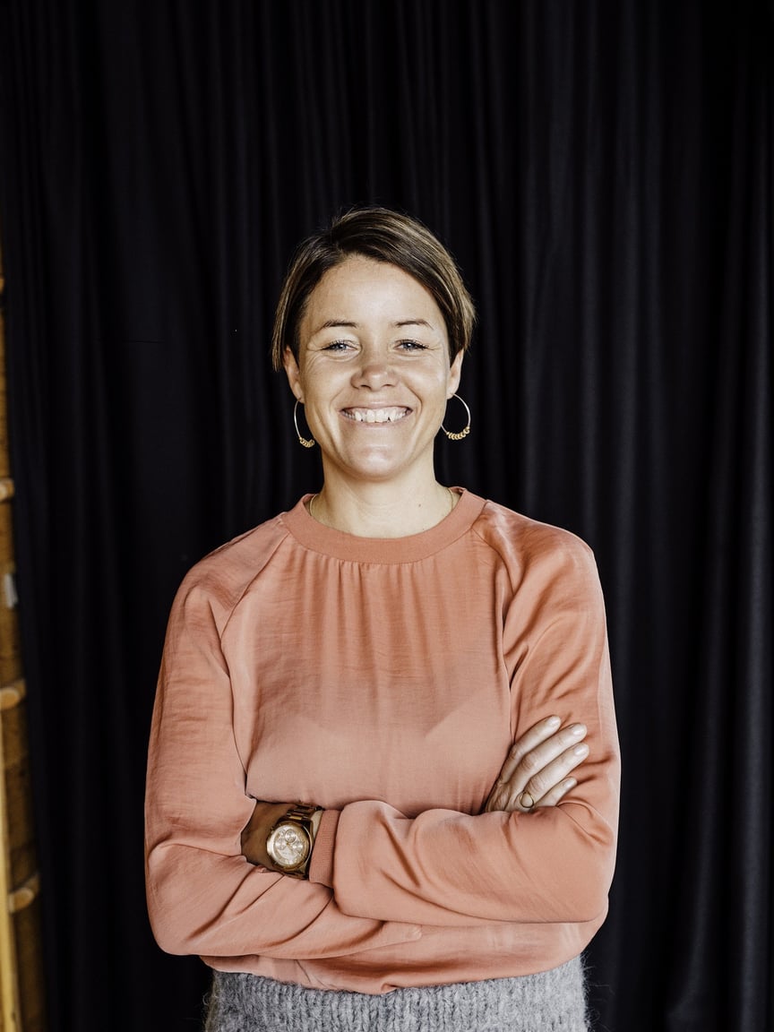 En smilende dame i rosa skjorte står foran en mørk bakgrunn med hendene i lommene og ser mot kamera.