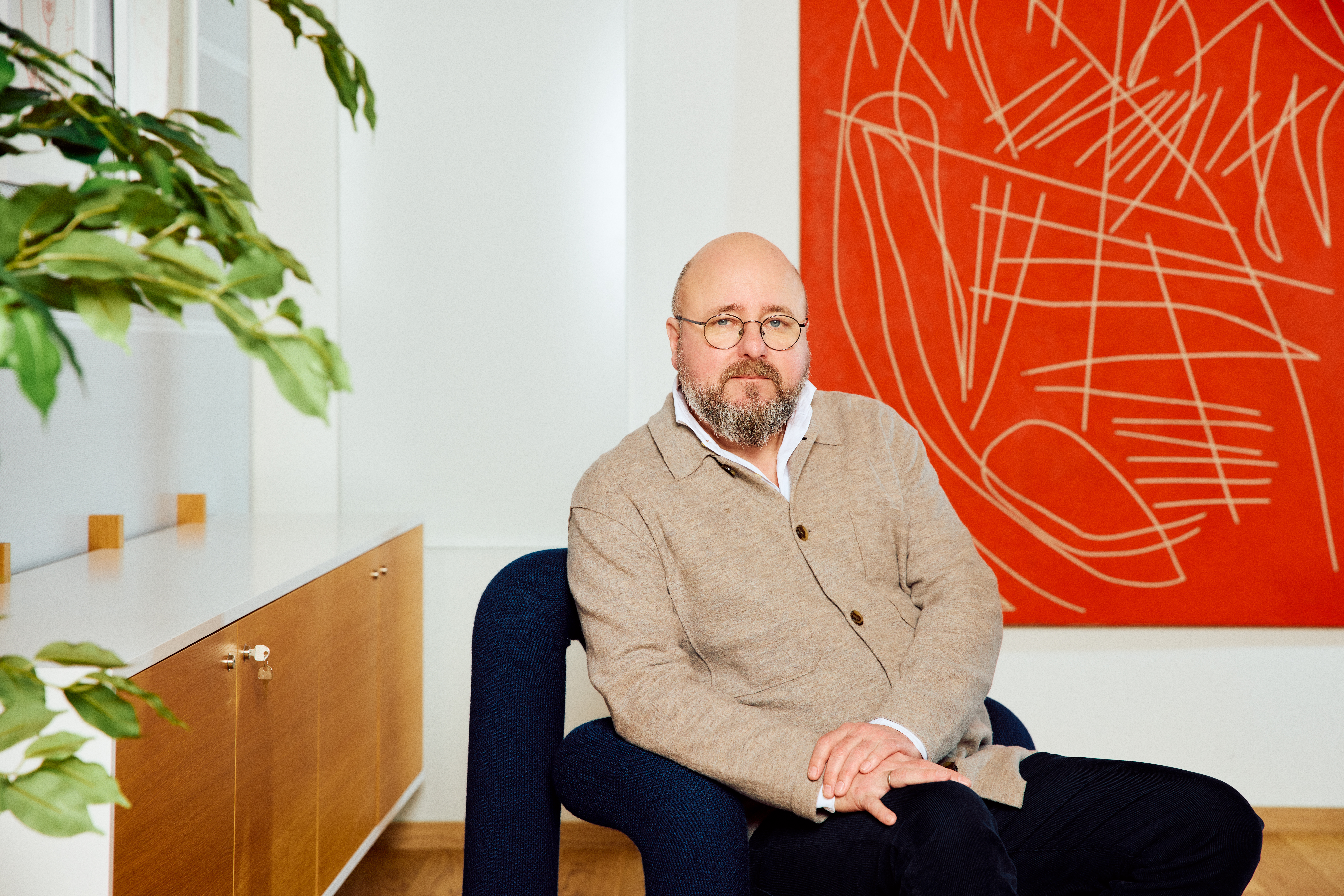 En mann sitter på en stol midt i bildet og ser rett mot kamera. Han har briller og en beige cardigan. I bakgrunnen står en plante, og et rødt kunstverk henger på veggen.