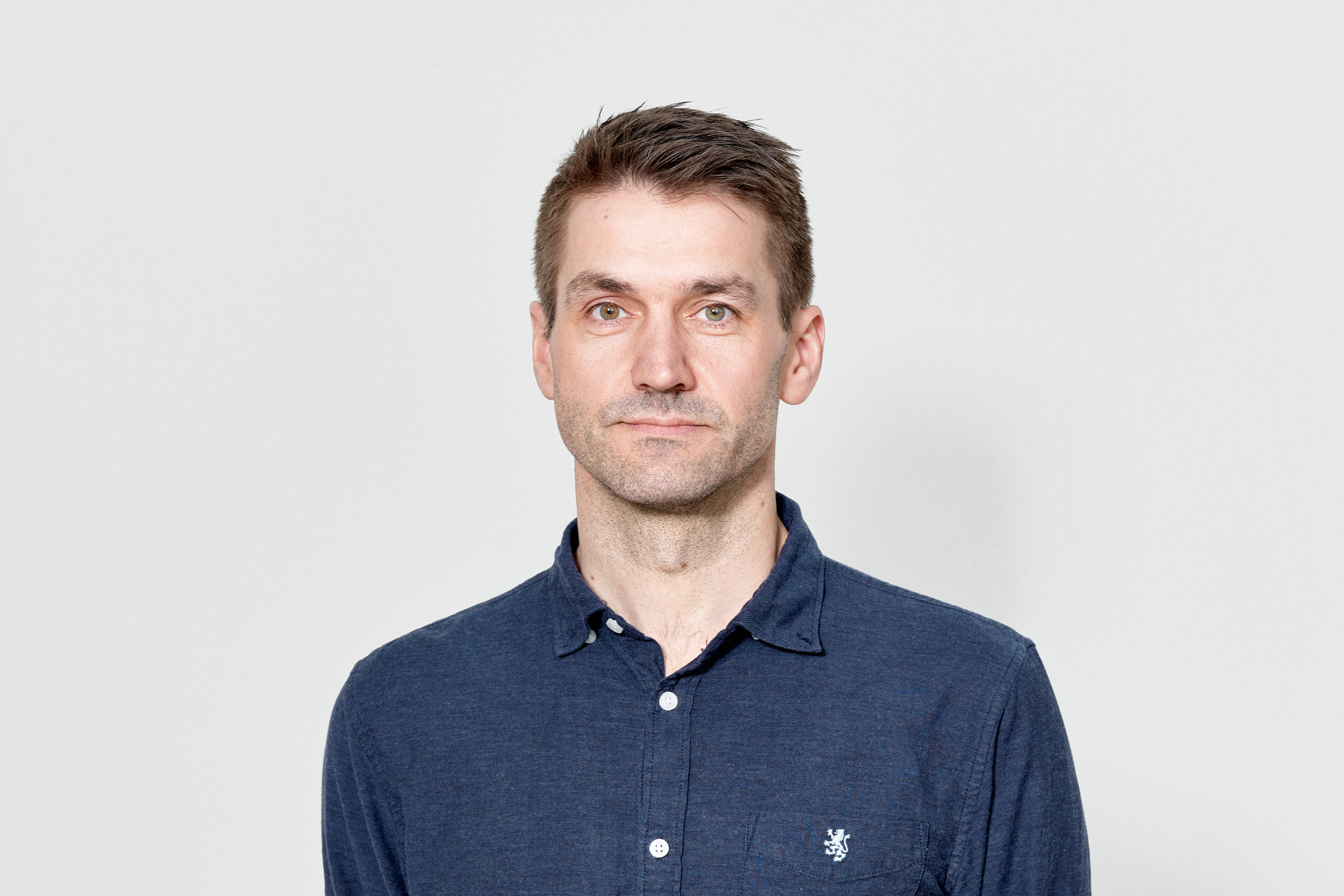 En mann står foran en hvit vegg, ser mot kamera, og har kort brunt hår og mørk blå skjorte.