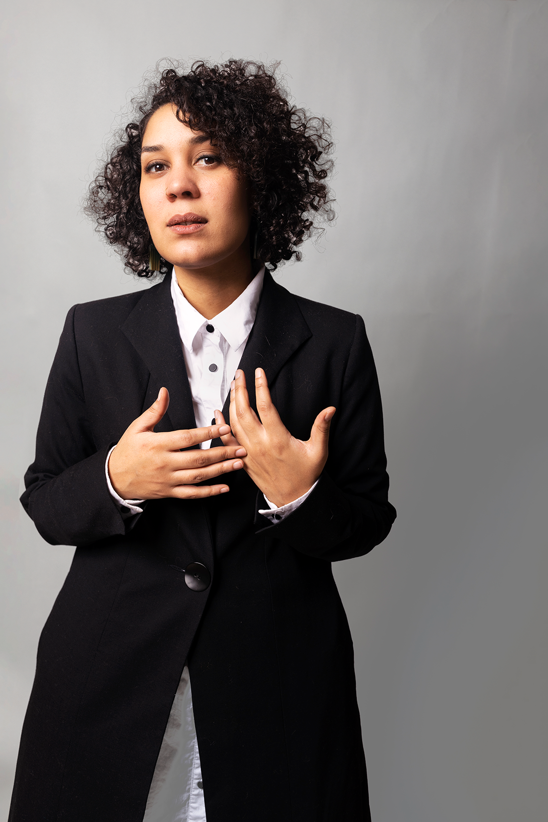 En kvinne står til venstre i bildet foran en grå vegg. Hun har brunt, krøllete hår og er kledd i en svart dressjakke med hvit skjorte under.