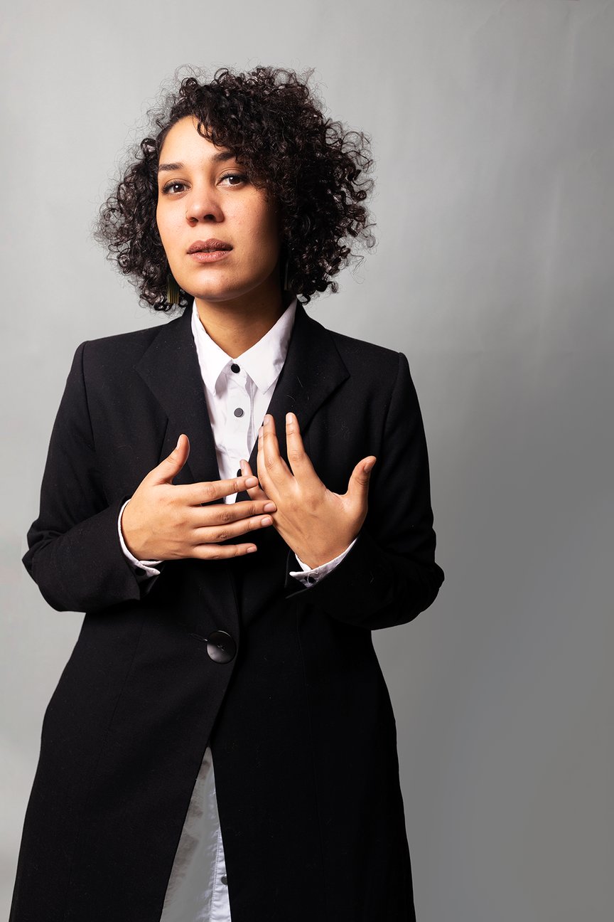 En kvinne står til venstre i bildet foran en grå vegg. Hun har brunt, krøllete hår og er kledd i en svart dressjakke med hvit skjorte under.