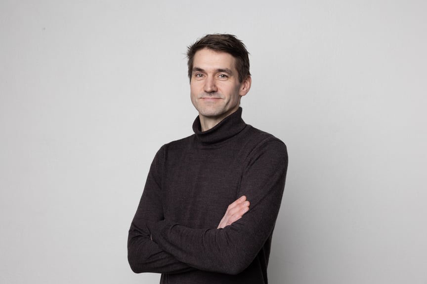 En mann står midt i bildet foran en hvit vegg. Han har kort brunt hår, svart høyhalset genser, smiler mot kamera og har armene krysset foran kroppen.