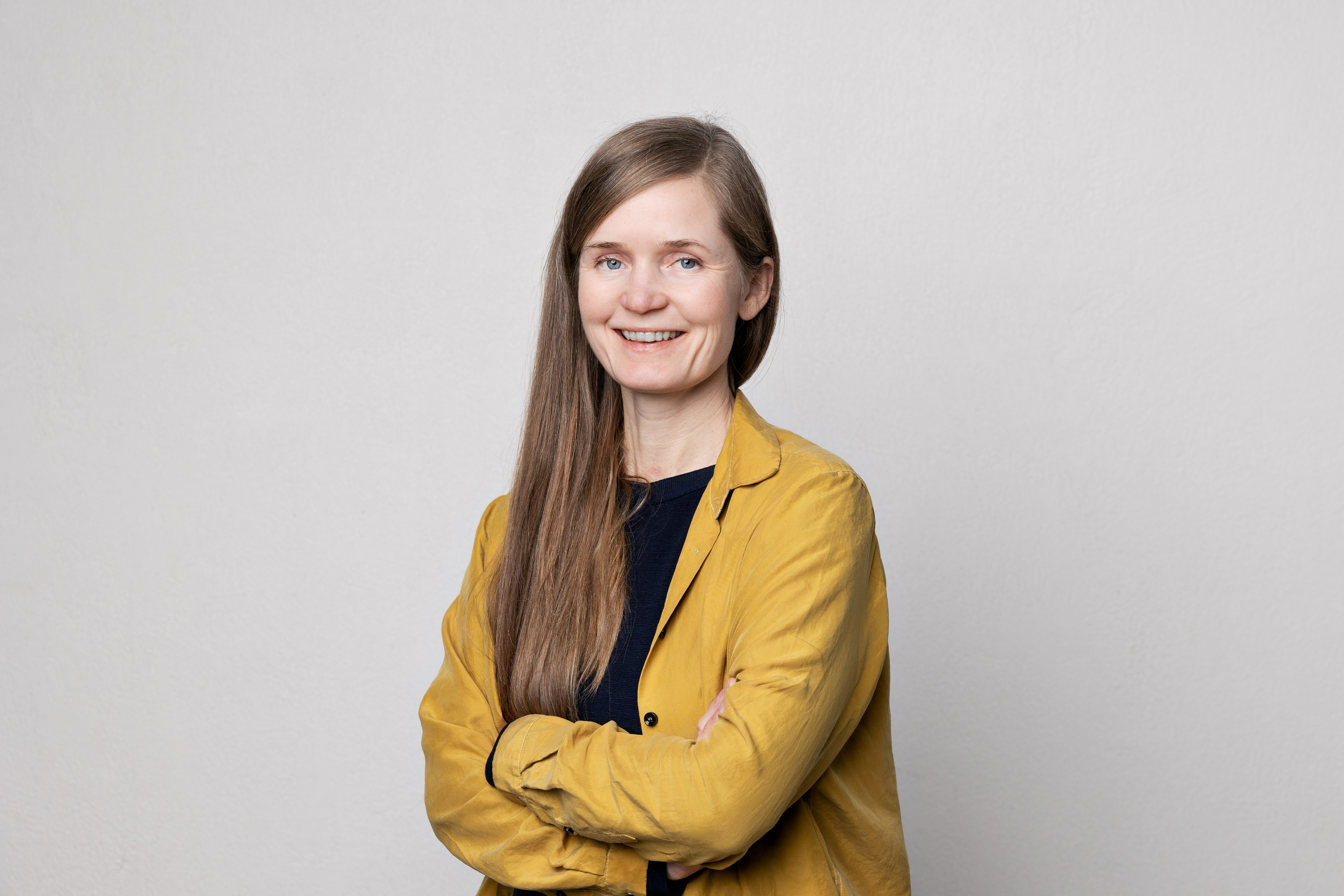 En kvinne med gul jakke og langt brunt hår står midt i bildet og smiler mot kamera