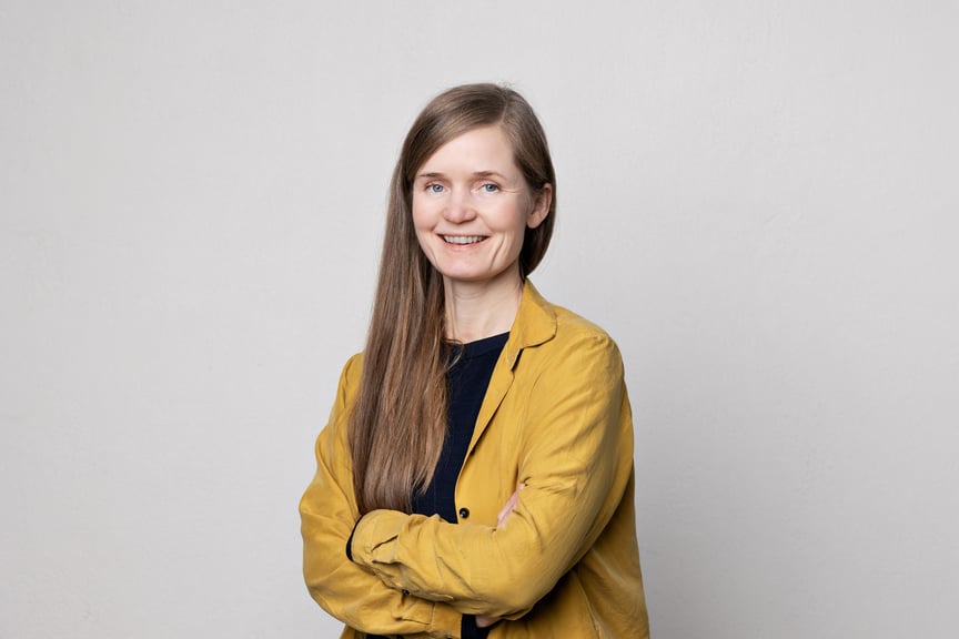 En kvinne med gul jakke og langt brunt hår står midt i bildet og smiler mot kamera