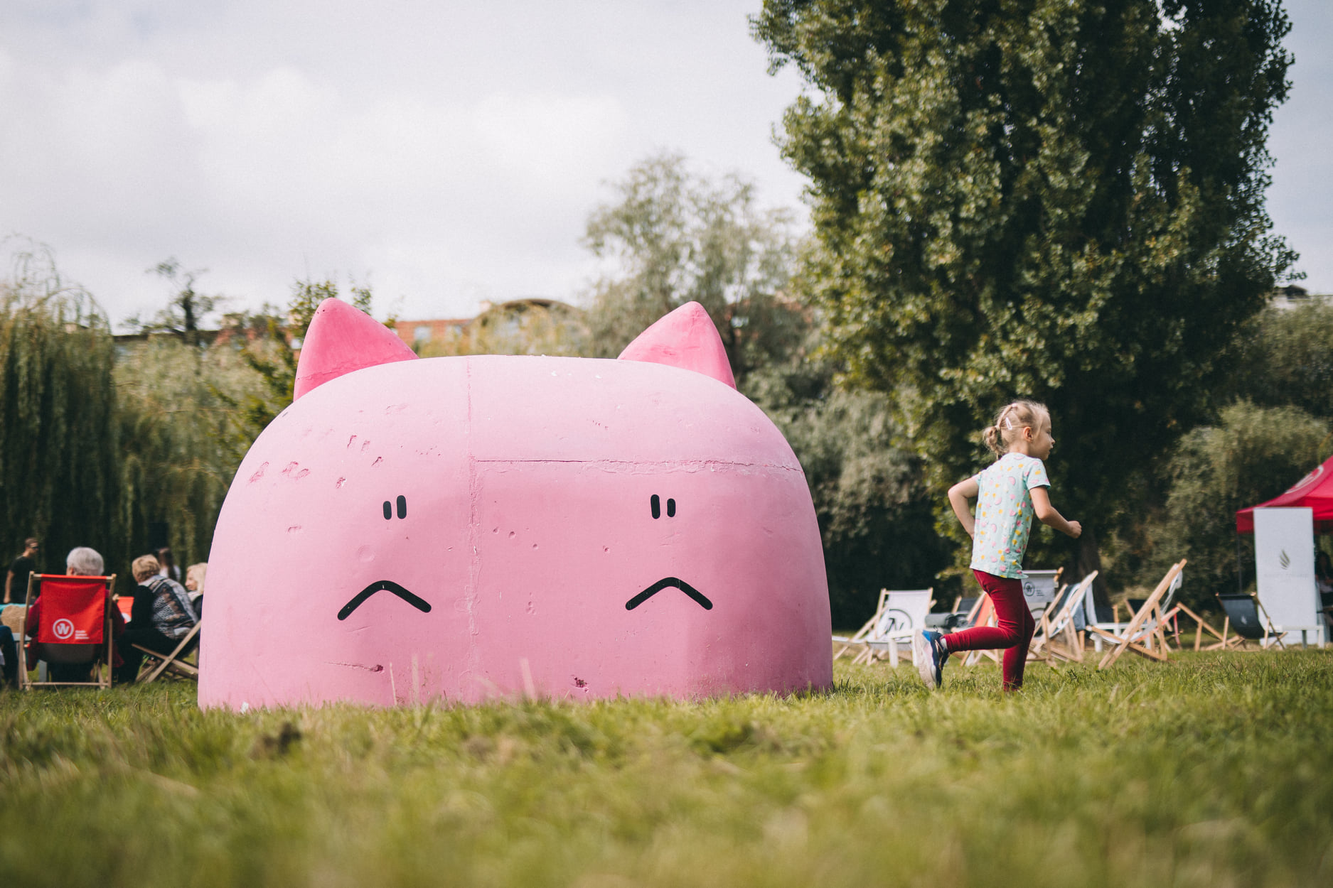 Stor rosa skulptur formet som et tegneserieaktig dyrehode med spisse ører står på en gresslette i en park. Et barn løper forbi på høyre side, mens flere personer sitter i stoler i bakgrunnen omgitt av trær og grøntområder.