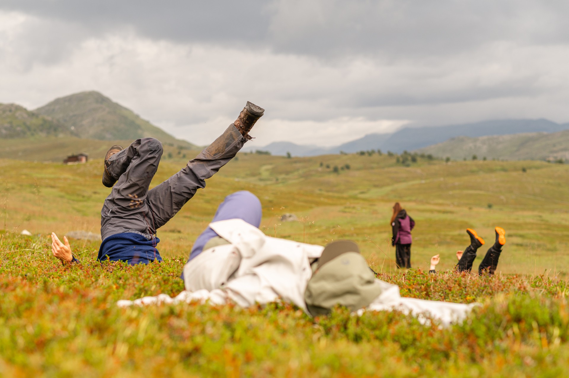 Bilde av dansere i et åpent høstgult landskap. Danserne har turklær på seg og ligger mot bakken i ulike positurer.