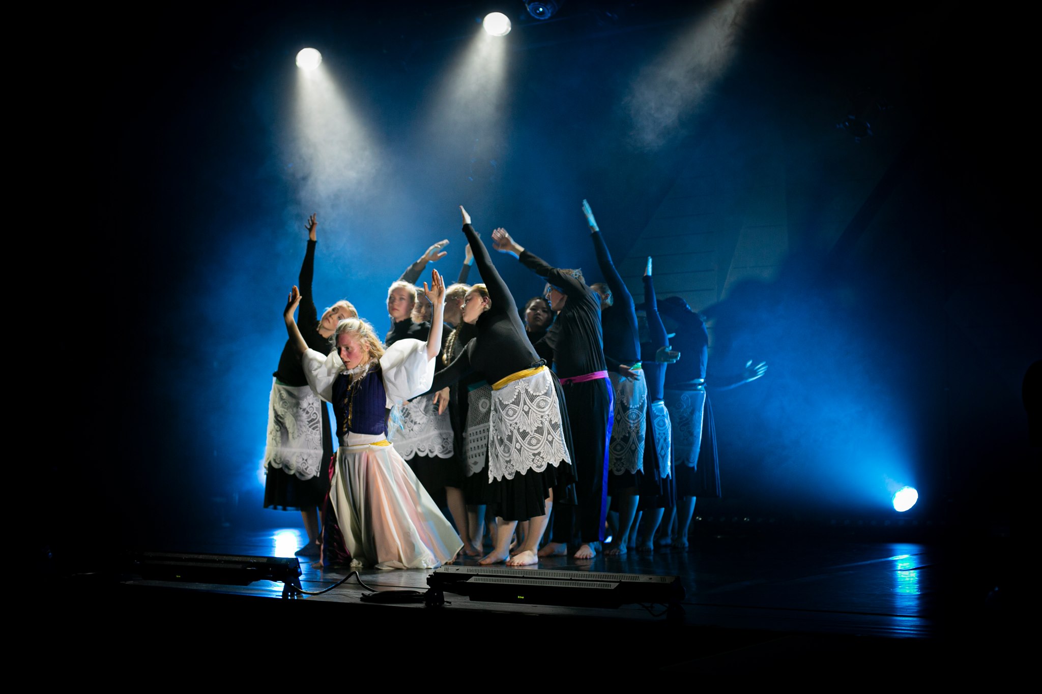 En gruppe dansere opptrer på en mørklagt scene under blått spotlys. Danserne står tett sammen i en koreografert formasjon med armene løftet og utstrakt i ulike retninger.