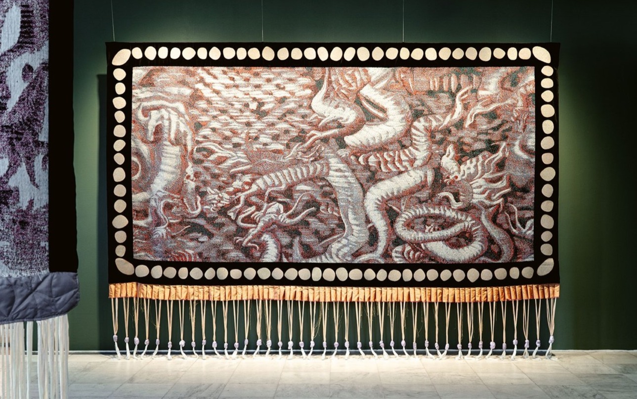 Et stort, detaljert veggteppe henger på en mørk grønn vegg. Motivet viser flere drager i rødt, hvitt og grått som slynger seg rundt hverandre i en dynamisk komposisjon. Rundt motivet går en dekorativ kant med hvite sirkulære mønstre. Nederst henger lange dusker av tau og tekstil, jevnt fordelt, som gir teksturen ekstra dybde. Teppeinstallasjonen er festet med synlige vaiere fra taket, og lyset i rommet fremhever både fargene og skyggene i verket.