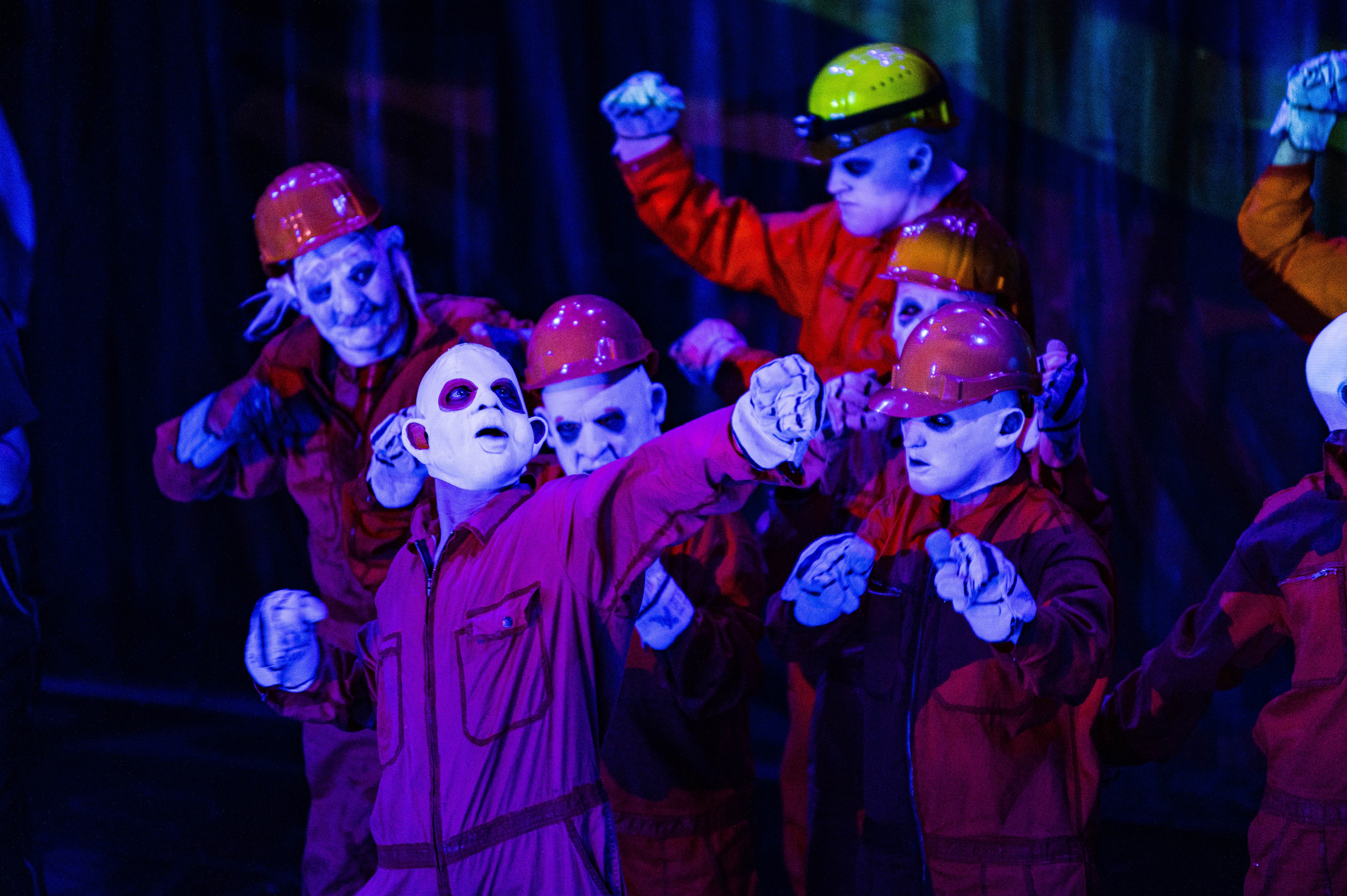 En gruppe personer i røde kjeledresser, hvite masker og fargerike hjelmer opptrer på en scene, belyst av sterkt blått lys.