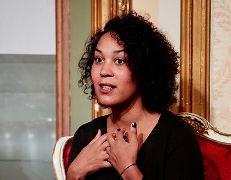 En person med krøllete hår sitter i en rød og gullfarget, ornamentert stol i et elegant interiør. Vedkommende har på seg en svart topp og holder hendene mot brystet, som om de uttrykker noe i samtale. Bakgrunnen består av en vegg med gullramme og en glassmonter, noe som gir rommet et sofistikert og historisk preg. Bildet formidler en personlig og uttrykksfull scene i et formelt miljø.