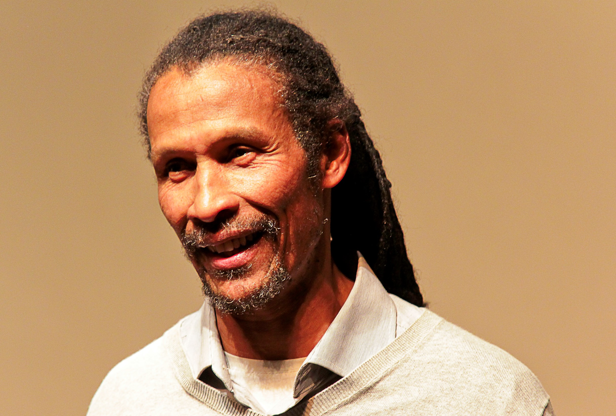 Portrett av en person med lange dreadlocks, iført en lys grå genser over en skjortekrage. Personen har et lett smil og et uttrykksfullt ansikt, som gir inntrykk av å være i samtale. Bakgrunnen er ensfarget, i beige eller lys brun tone, og gir et rolig og nøytralt visuelt uttrykk.