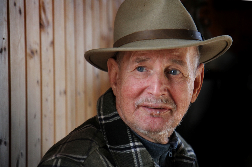 En eldre person med lys hud, blå øyne og kort grått skjegg står foran en vegg med loddrette trepaneler. Vedkommende har på seg en beige hatt i fedora-stil og en rutete jakke, og ser rolig til siden med nøytralt uttrykk. Bildet har varme toner og tydelige teksturer i både klær og bakgrunn, og formidler et portrett med karakter og tilstedeværelse i et rustikt miljø.