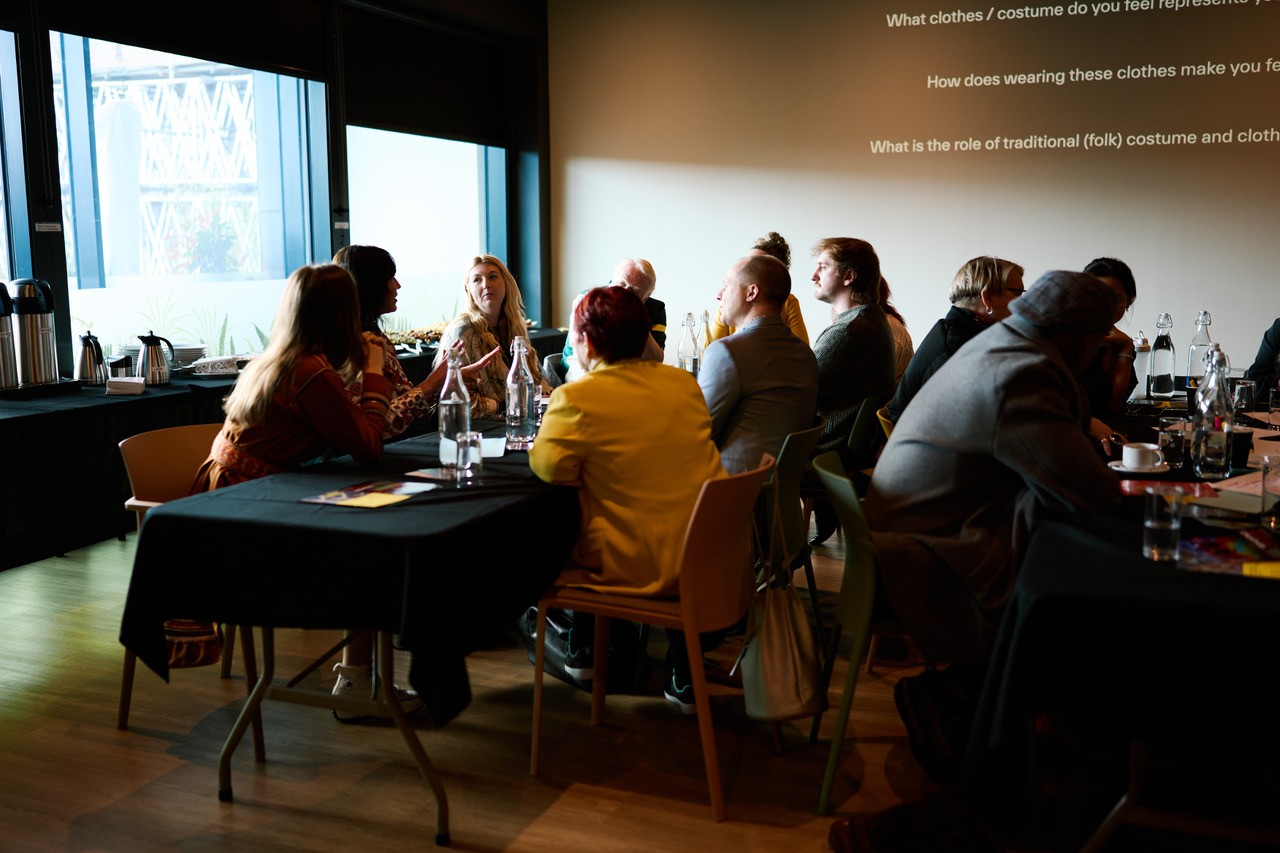 En gruppe mennesker sitter rundt bord i et dempet belyst rom, trolig under en workshop eller diskusjonsøkt. Store vinduer slipper inn naturlig lys fra én side, og en prosjektor viser spørsmål på veggen om klær og kulturell identitet. Deltakerne virker engasjerte, med vannflasker, papirer og glass på bordene, noe som tyder på en organisert og reflekterende samling.