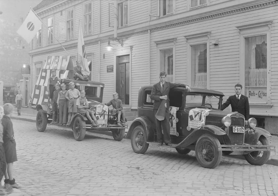 Sort-hvitt bilde fra en gate i en eldre by, sannsynligvis fra 1930- eller 40-tallet. To gamle biler står parkert ved brosteinsveien foran et trehus. Den første bilen har flere personer, inkludert barn, på en dekorert tilhenger med flagg og bannere. Den andre bilen har to unge menn som står foran og ved siden av bilen. Det er flere tilskuere, blant annet barn, som ser på. Både biler og mennesker er pyntet til en slags feiring eller parade.