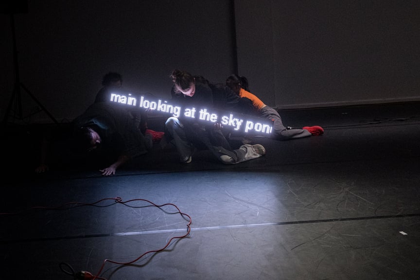 Bildet viser fire dansere som beveger seg på gulvet med en lysende tekstfremviser.