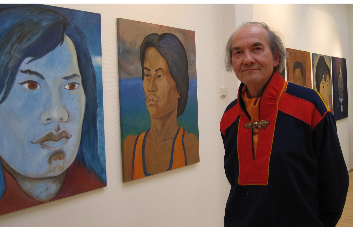 En mann står i et kunstgalleri ved siden av en rekke portrettmalerier som henger på en hvit vegg. Han har på seg en mørk jakke med røde og gule detaljer og en dekorativ lås foran. Maleriene viser stiliserte ansikter med ulike uttrykk og bakgrunner, malt med kraftige farger og penselstrøk. Rommet er godt opplyst, og bildet formidler et øyeblikk av kunstnerisk nærvær og visuell refleksjon, muligens med kunstneren selv foran sitt eget verk.