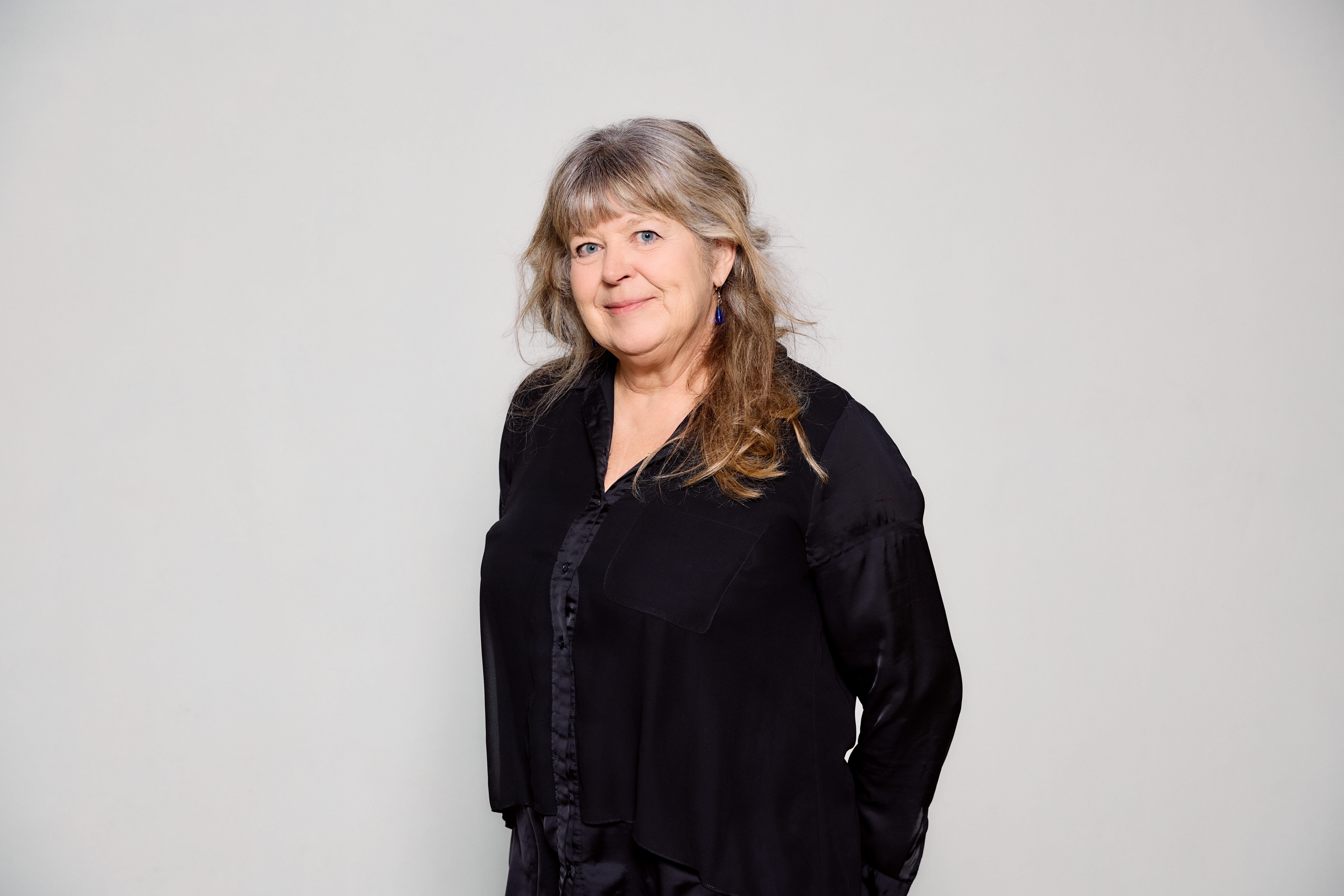 En kvinne med svart genser står midt i bildet foran en hvit vegg.