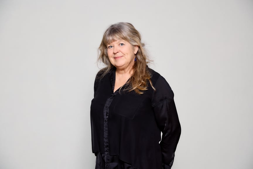 En kvinne med svart genser står midt i bildet foran en hvit vegg.