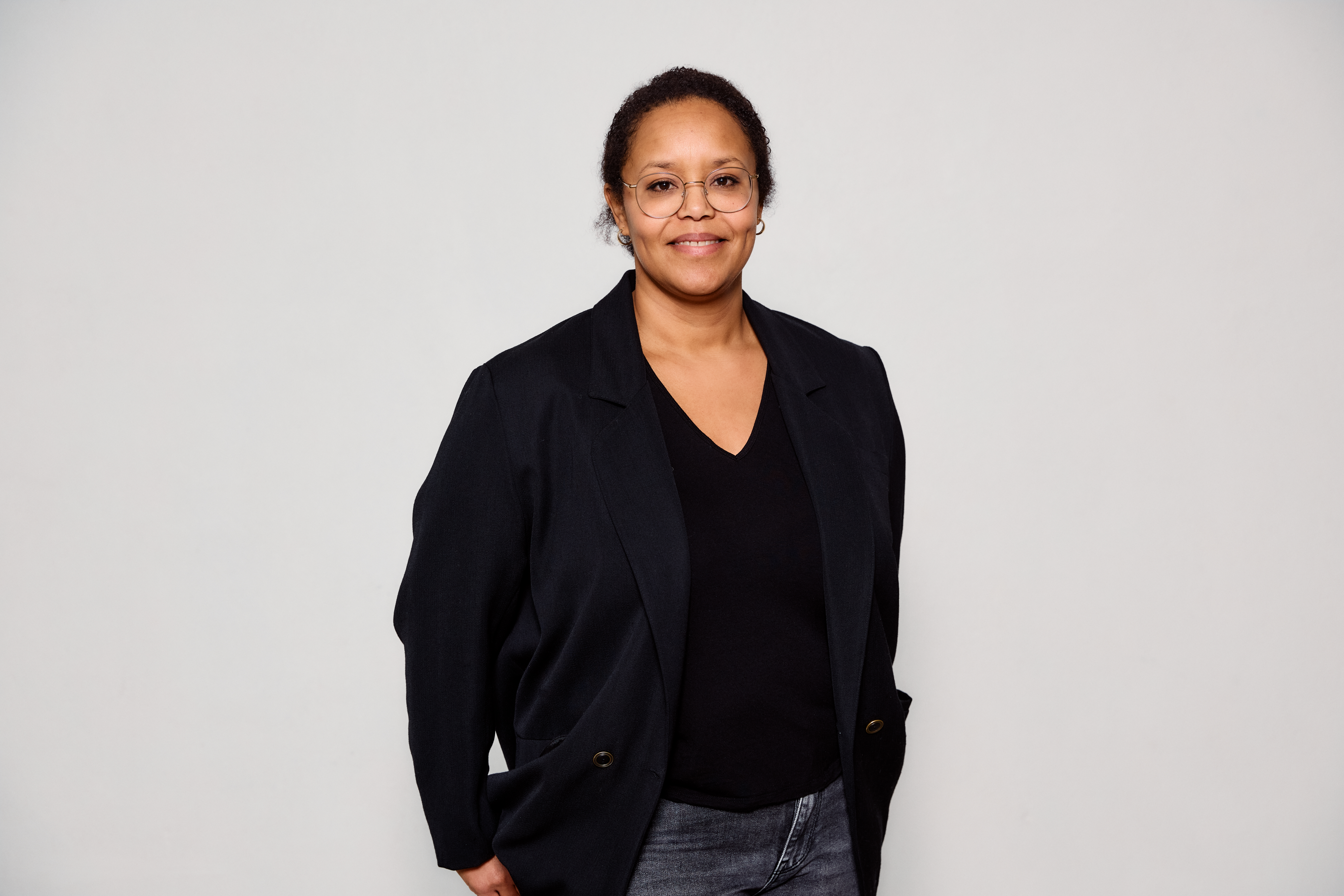 En kvinne med briller og svarte klær på overkroppen står midt i bildet foran en hvit vegg.