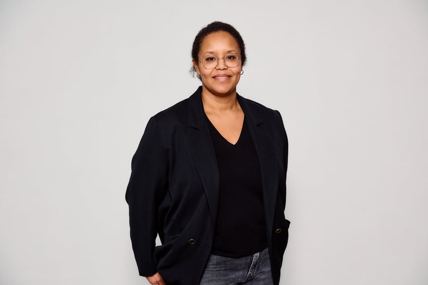 En kvinne med briller og svarte klær på overkroppen står midt i bildet foran en hvit vegg.