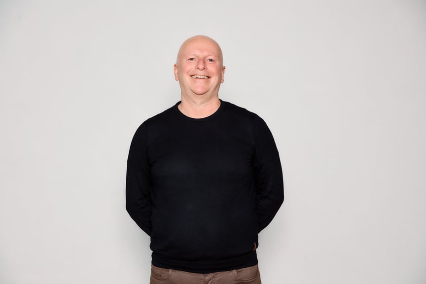 En mann med svart genser står midt i bildet foran en hvit vegg.