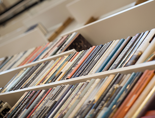 Flere hvite bokhyller står på rekke og rad, fylt med bøker i ulike farger og størrelser. Bøkene er plassert vertikalt med ryggene vendt utover, og fotografiet er tatt i vinkel som fremhever dybden og den ryddige organiseringen. Bildet viser et lyst og strukturert bibliotek- eller bokhandelmiljø, og formidler ro, orden og mangfold i litteratur.