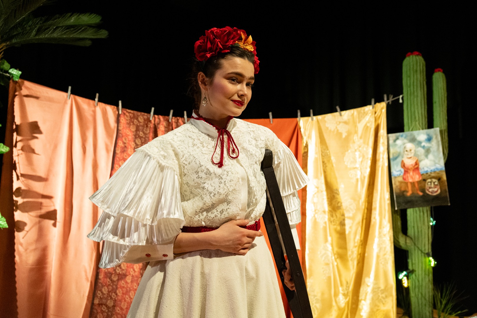 En kvinne i kostyme inspirert av Frida Kahlo står på en scene under en forestilling. Hun har fargerike klær, blomster i håret og markerte øyenbryn, og ser ut mot publikum i dramatisk scenebelysning.