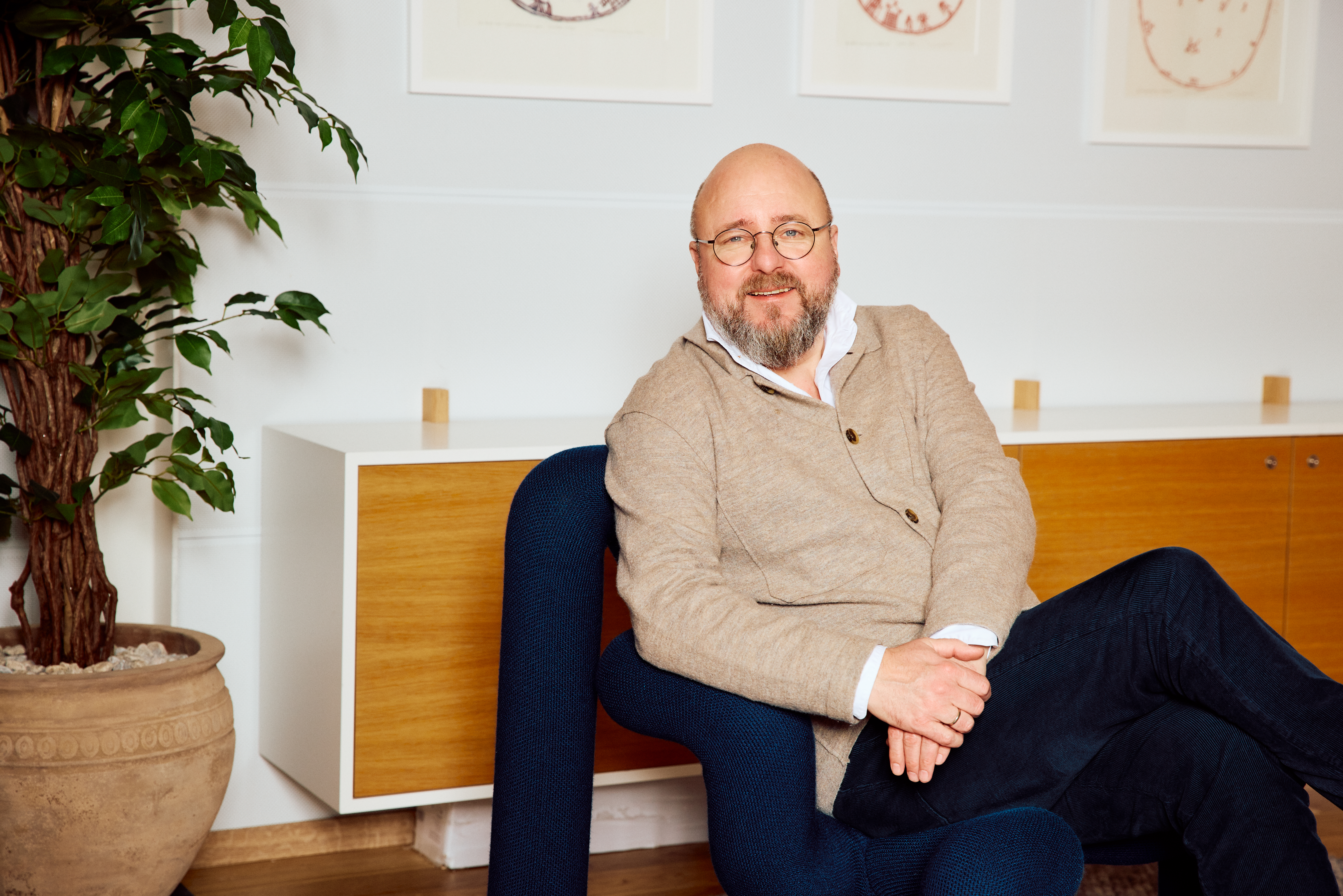En mann sitter i en stol i et møblert rom med beina i kryss og smiler mot kamera.