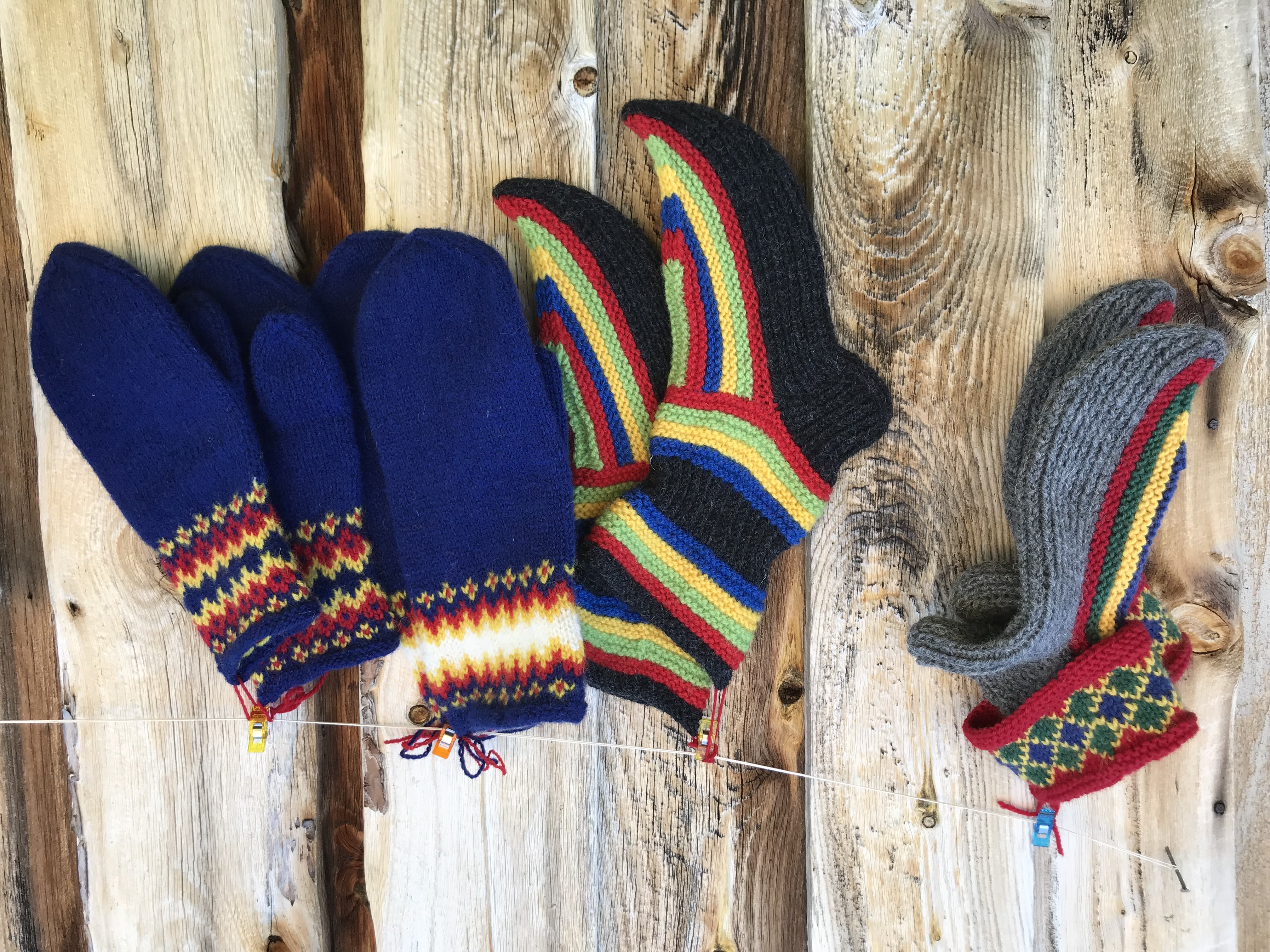 Seks fargerike, strikkede sokker og votter henger på en tynn snor med små klesklyper i ulike farger. Plaggene har livlige mønstre i rødt, gult, grønt, blått og svart, og er arrangert foran en bakgrunn av loddrette, værbitte treplanker. Bildet gir en varm og tradisjonell følelse, og fremhever håndverk, tekstur og fargerik kultur i et rustikt miljø.