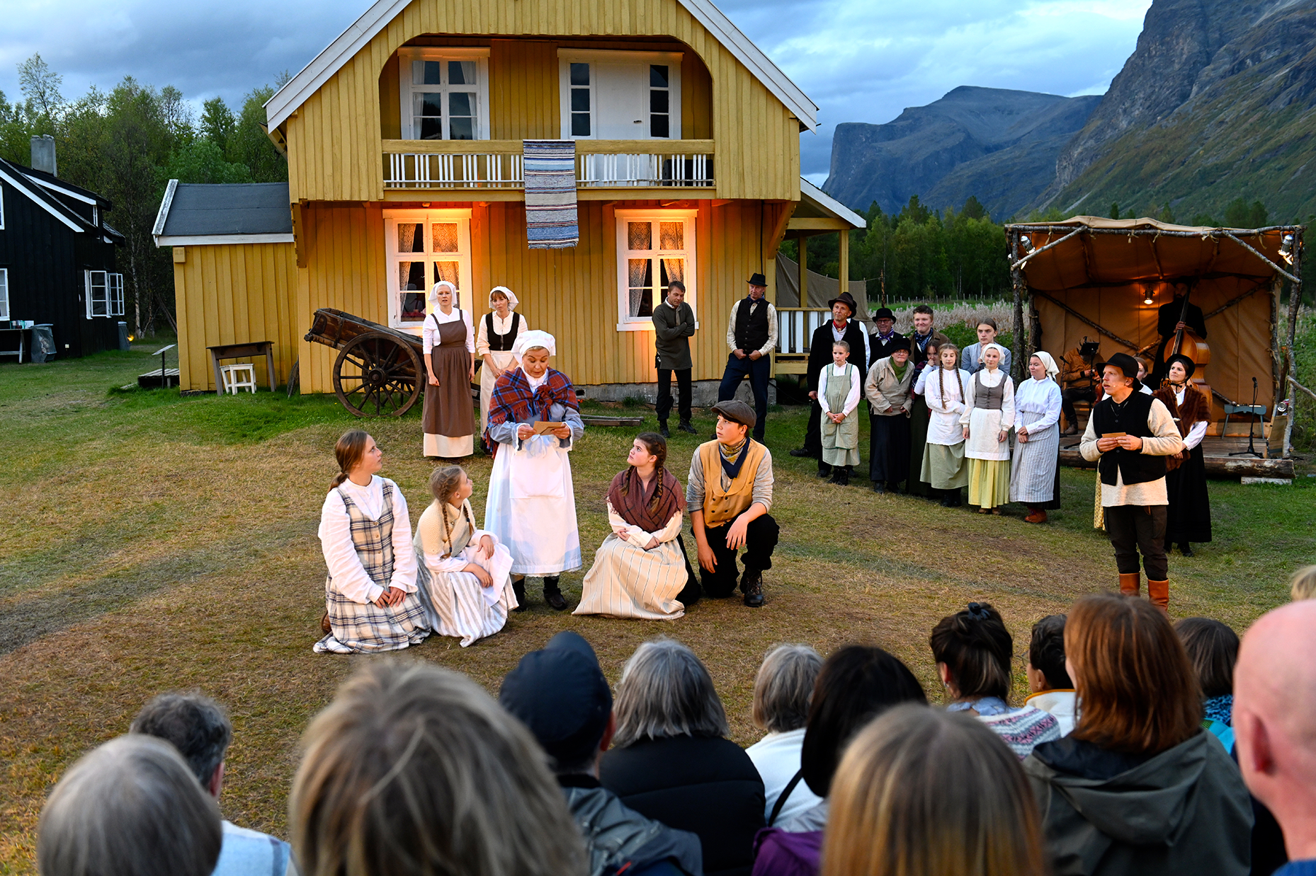 Friluftsteater foran et gult trehus i landlige omgivelser. En gruppe skuespillere i tradisjonelle, historiske kostymer opptrer foran et publikum som sitter utendørs. I bakgrunnen er det fjell og skog, og stemningen er varm med lys fra huset og en scene på høyre side.