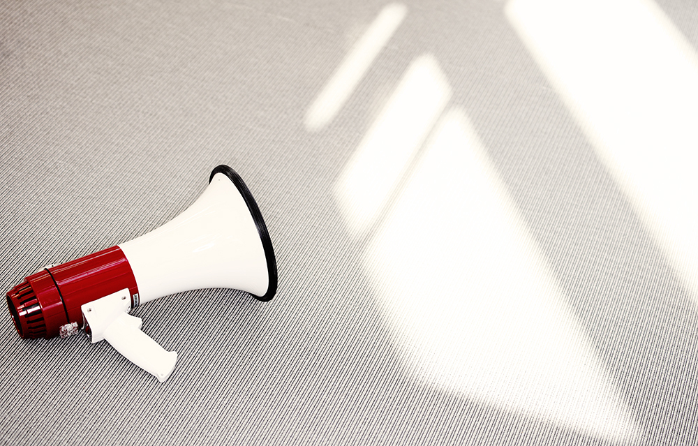 En rød og hvit megafon ligger på et lyst, teksturert underlag, trolig et teppe eller en matte. Megafonen er plassert mot venstre side av bildet, med munnstykket vendt mot venstre og åpningen mot høyre. Fra øvre høyre hjørne strømmer geometriske lysflekker inn, som skaper skarpe kontraster mellom lys og skygge på underlaget. Bildet formidler et stille øyeblikk med potensial for kommunikasjon, og har et estetisk preg med fokus på lys, form og symbolikk.