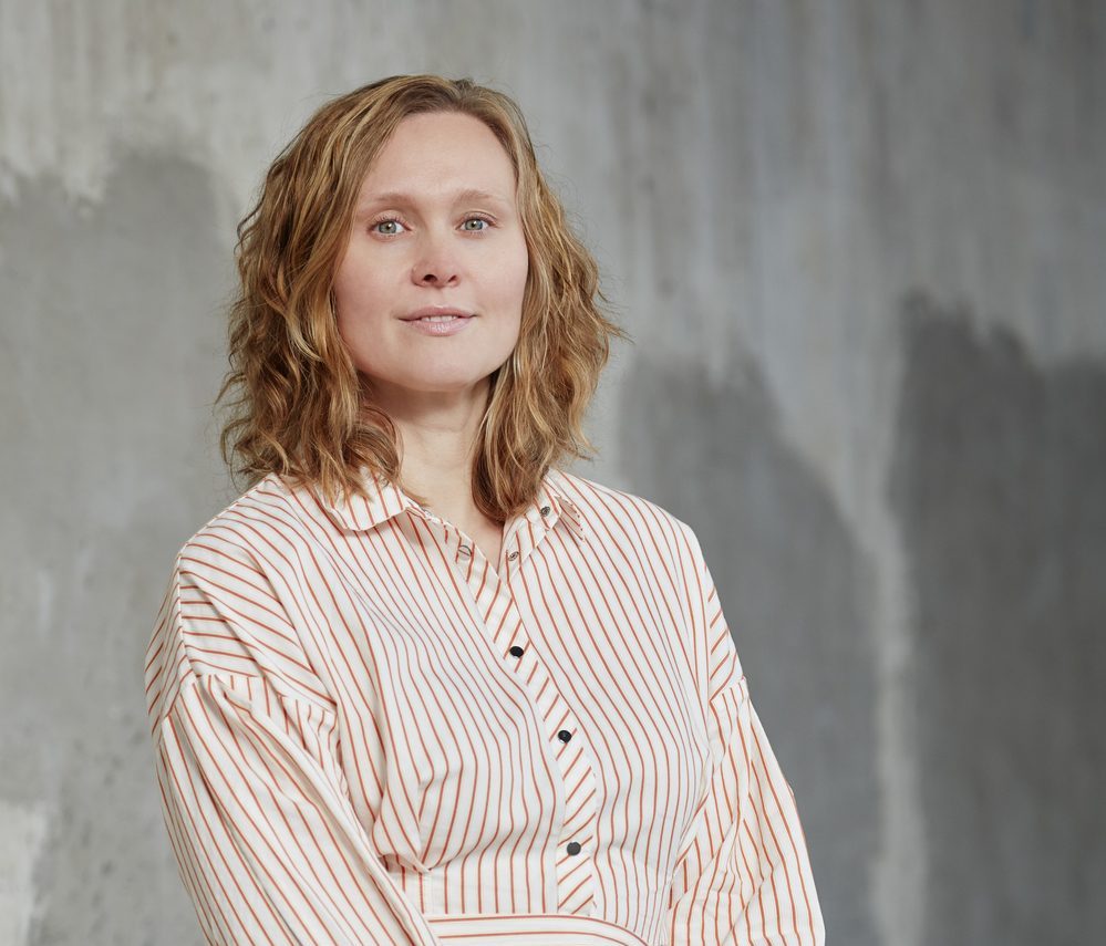 En person med skulderlangt, bølget hår står foran en vegg med grå, teksturert overflate. Vedkommende har på seg en lang, hvit kjole med røde striper i vertikale og diagonale mønstre, belte i midjen og ermer til albuene. Personen står med hendene samlet foran seg og ser rett mot kameraet. Den industrielle bakgrunnen skaper kontrast til det ryddige og grafiske antrekket, og bildet formidler ro, styrke og visuell balanse.