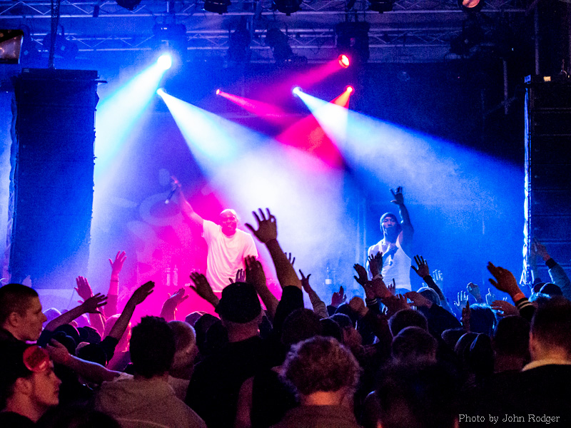 Publikum med hevede hender deltar entusiastisk på en konsert eller musikkarrangement. To artister står på scenen, badet i fargerikt scenelys i blått, rødt og lilla, og én av dem peker mot publikum. Stemningen er energisk og festlig, med tettpakket folkemengde og profesjonelt lys- og lydutstyr som rammer inn opplevelsen. Bildet fanger et levende øyeblikk av musikkglede og fellesskap.