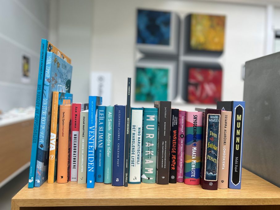 Et bilde av en trehylle med bøker stående tett i rekke. Bøkene er organisert side ved side med ryggen vendt ut, og varierer i farger og størrelser. I bakgrunnen sees et moderne interiør med abstrakt kunst på veggen og en disk eller benk til venstre i bildet. Atmosfæren gir inntrykk av et bibliotek, en bokhandel eller et personlig lesehjørne.