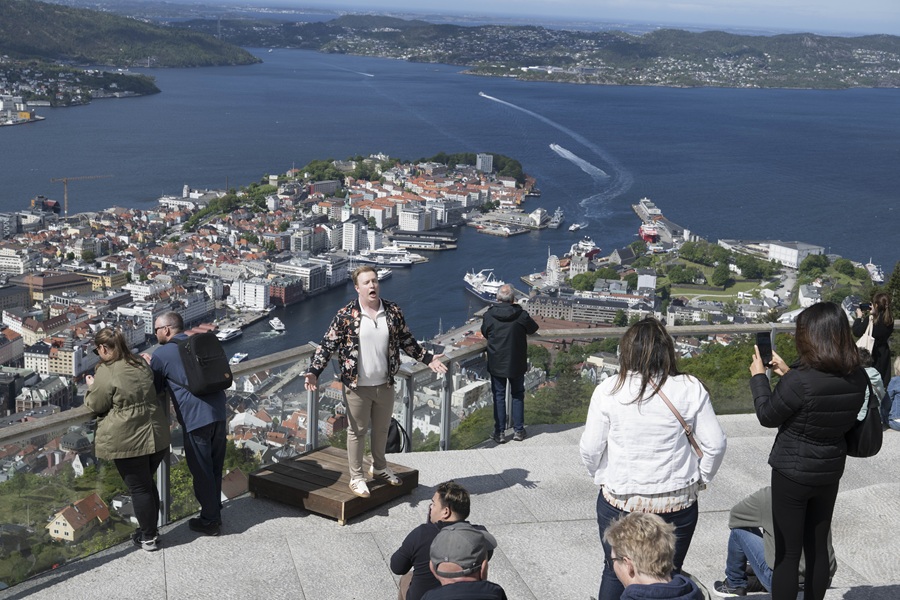 Flere personer står eller sitter på en utsiktsplattform med panoramautsikt over byen Bergen. Én person poserer på en liten forhøyning, mens andre tar bilder eller nyter utsikten. I bakgrunnen sees tettbygde hus, havneområdet og fjorder omgitt av fjell. En båt lager tydelige bølger i vannet, og landskapet viser en blanding av urban struktur og storslått natur. Bildet formidler turisme, utforskning og naturopplevelse i en norsk kystby.