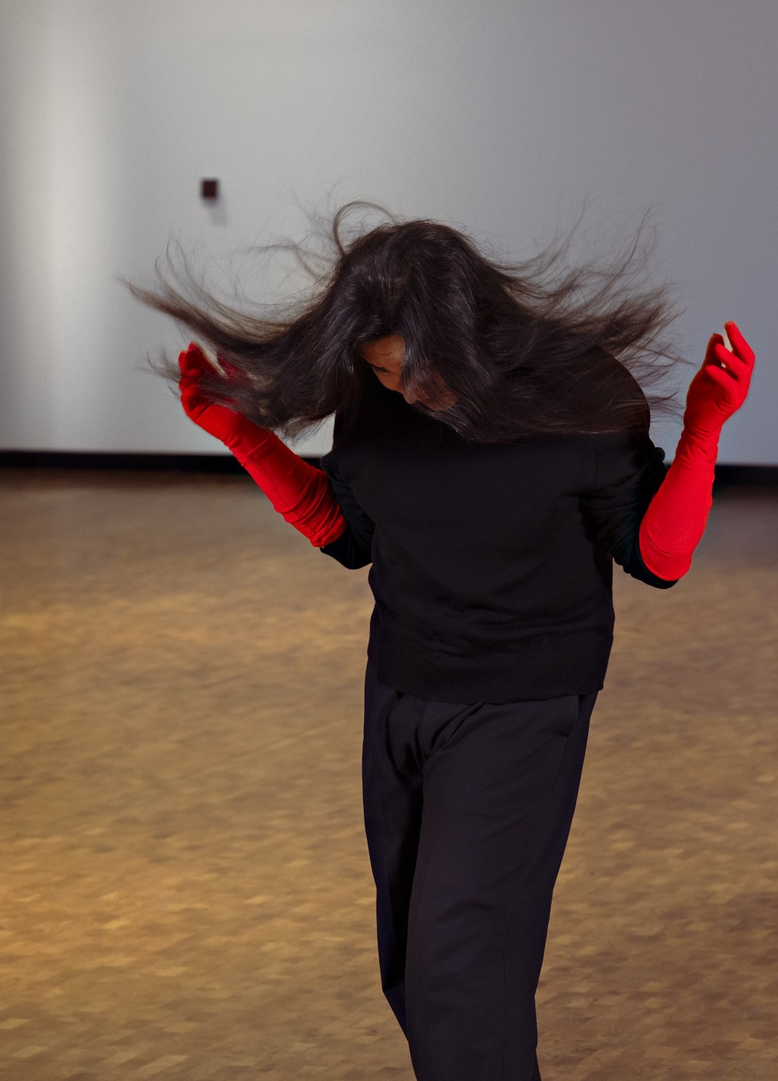 En person står oppreist og ser mot gulvet, kledd i svart, med hendene i en dansende positur.