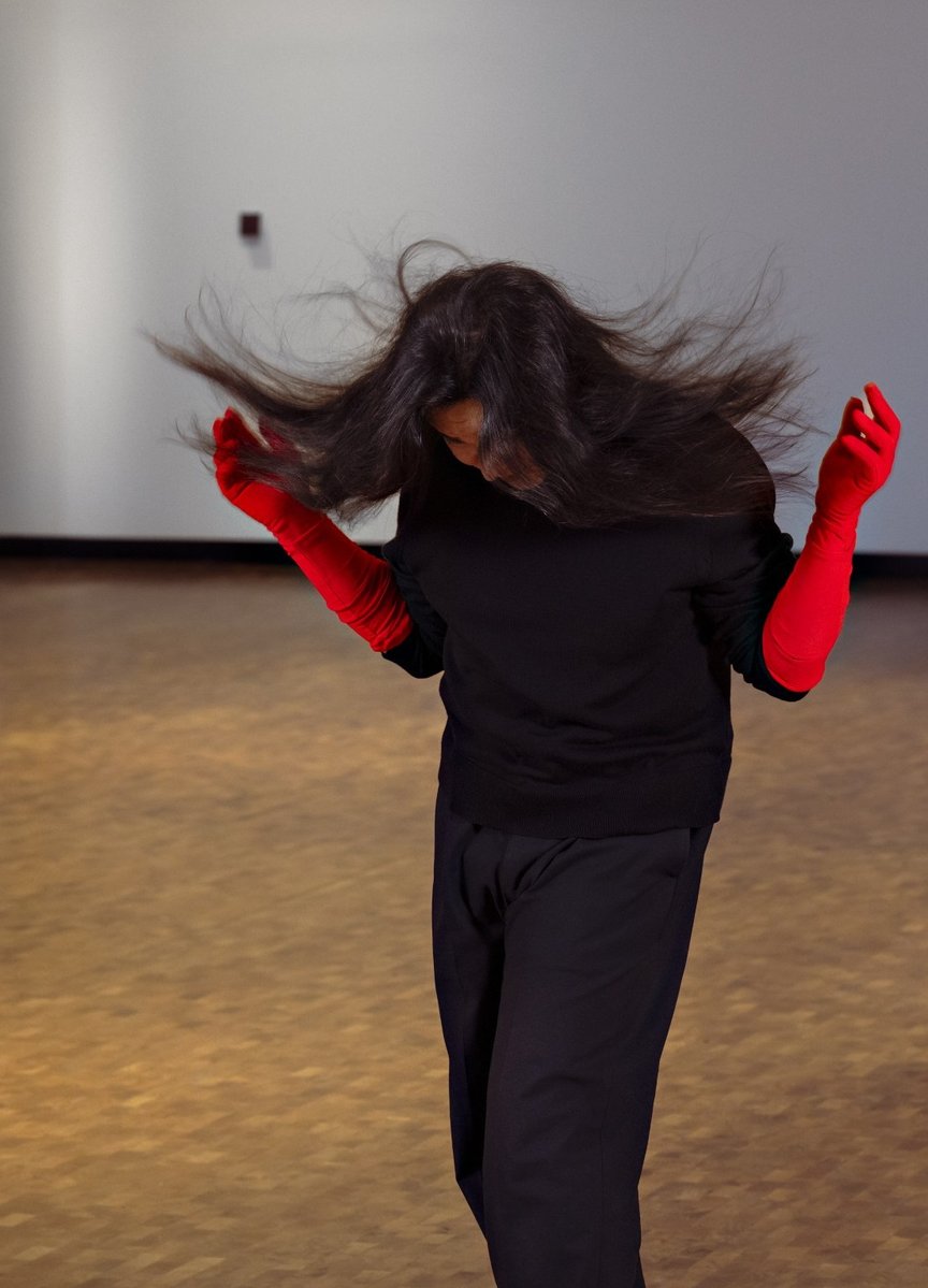 En person står oppreist og ser mot gulvet, kledd i svart, med hendene i en dansende positur.