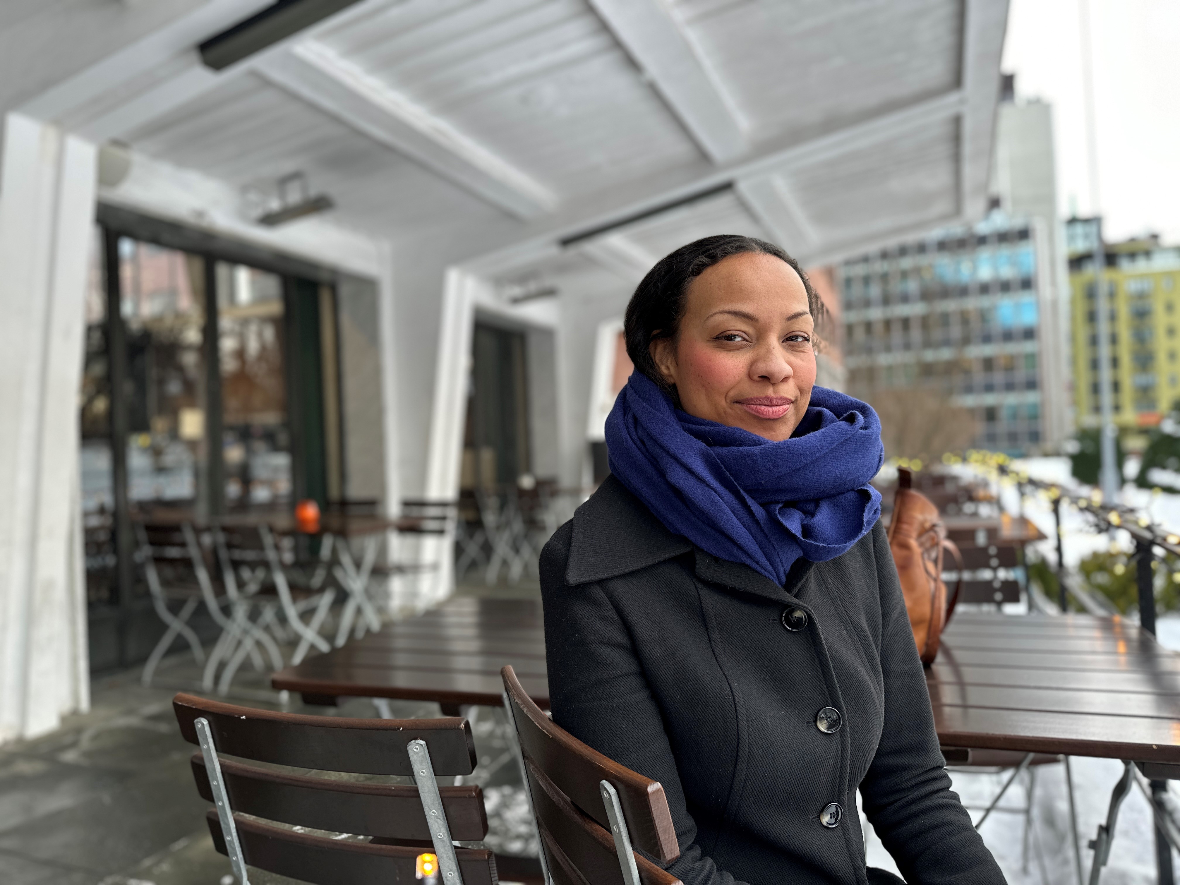 En person med mørk frakk og blått skjerf sitter på en trestol med metallben ved et bord i et utendørs serveringsområde. Området har flere bord og stoler arrangert i rekker under en hvit overbygning. I bakgrunnen sees bygninger og trær pyntet med lys, som antyder vinterstemning. Bakken er våt, trolig etter regn eller smeltet snø. Bildet formidler en rolig urban atmosfære med kontrast mellom den varme påkledningen og det moderne, åpne kafémiljøet.